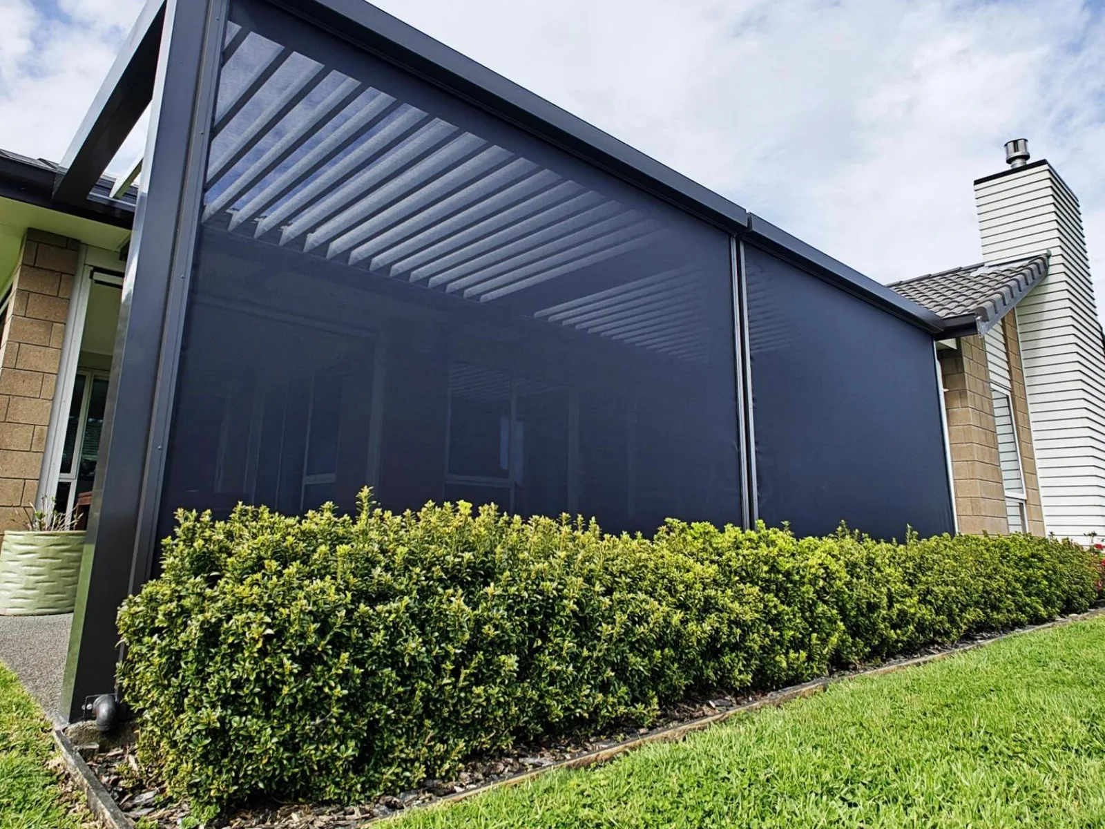 Dark mesh EasyTrack blinds closing in a louvre-roofed outdoor room behind a hedge